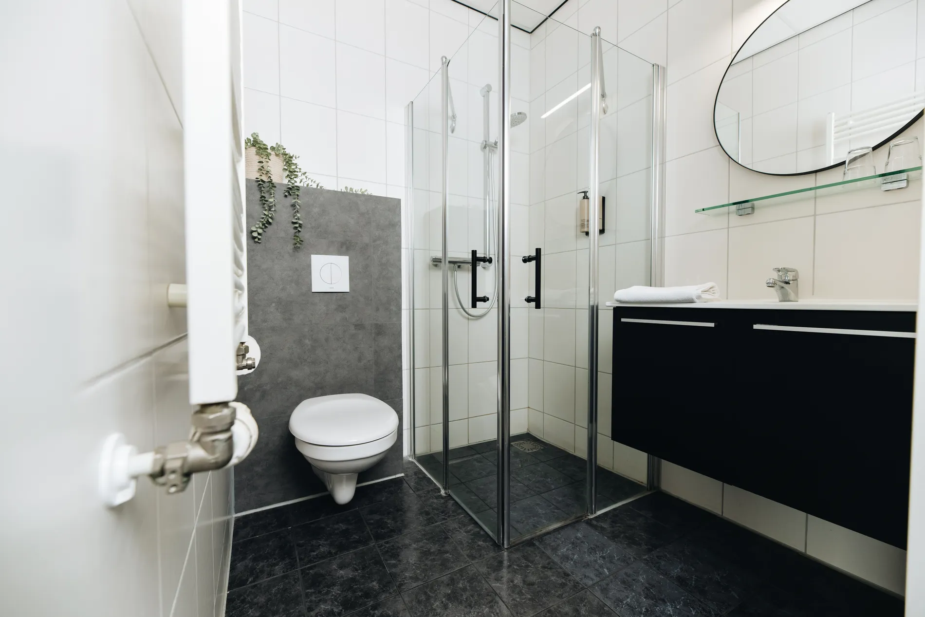 Moderne badkamer met glazen inloopdouche, zwart wastafelmeubel, ronde spiegel en wit-grijze betegeling.